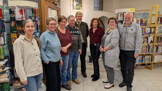 A group of smiling adults stand together in a room, possibly a library, wearing casual attire. They are looking at the camera.