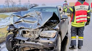 A severely damaged BMW car with a crumpled front end is on the side of the road, with a fireman nearby.