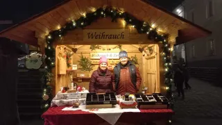 Two people stand in front of a decorated booth selling items. The booth has the sign 'Weihrauch'. Behind them, two people walk in the distance.