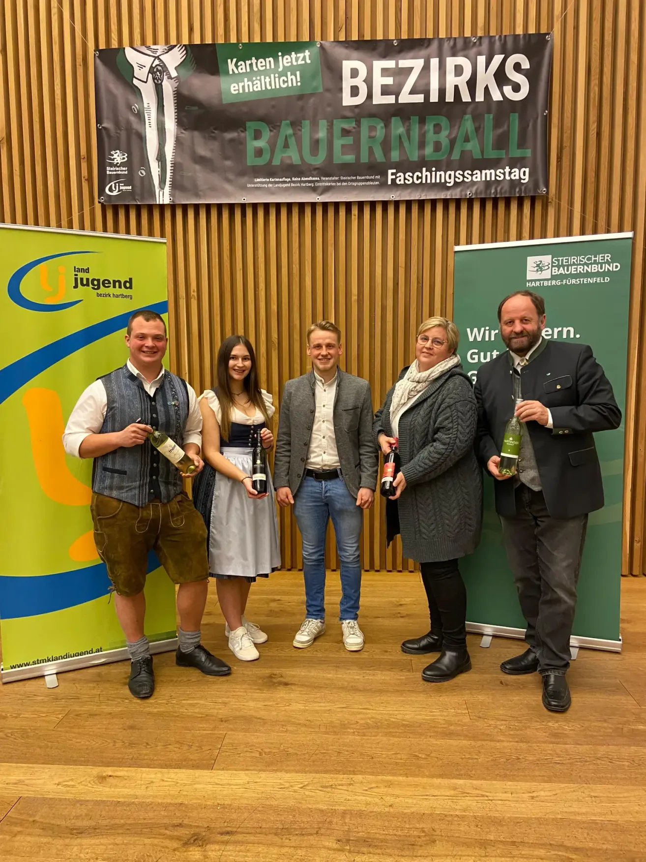 Five people in traditional German attire stand in front of a wooden wall, smiling for a photo. They hold bottles of beer and wine. Banners for 'Bauernball' and 'Steirischer Bauernbund' are behind them.