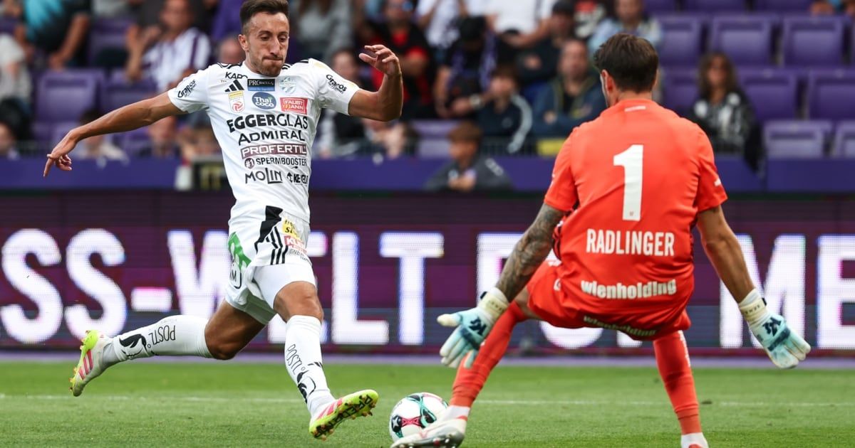 Soccer player in white jersey runs towards goal, while another player in red blocks with gloves on. Large crowd in background.
