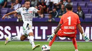 Soccer player in white jersey runs towards goal, while another player in red blocks with gloves on. Large crowd in background.