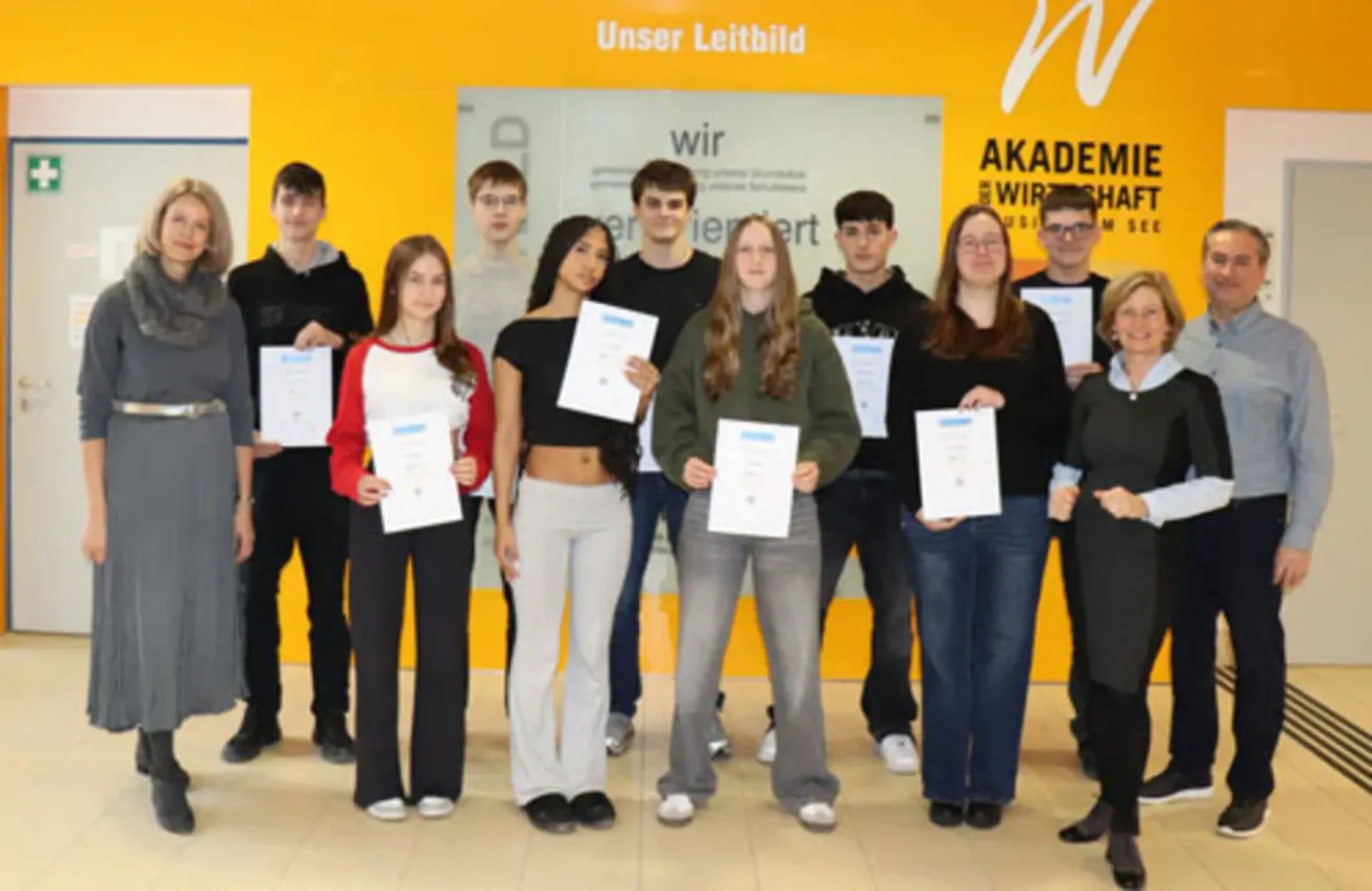 A group of young students in a school corridor holding certificates, standing in front of a yellow wall with text.
