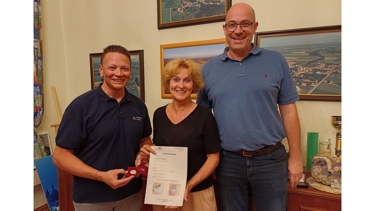 Three people stand in a room, smiling for a photo. A woman holds a paper certificate and a medal. Behind them, three framed pictures hang on the wall.