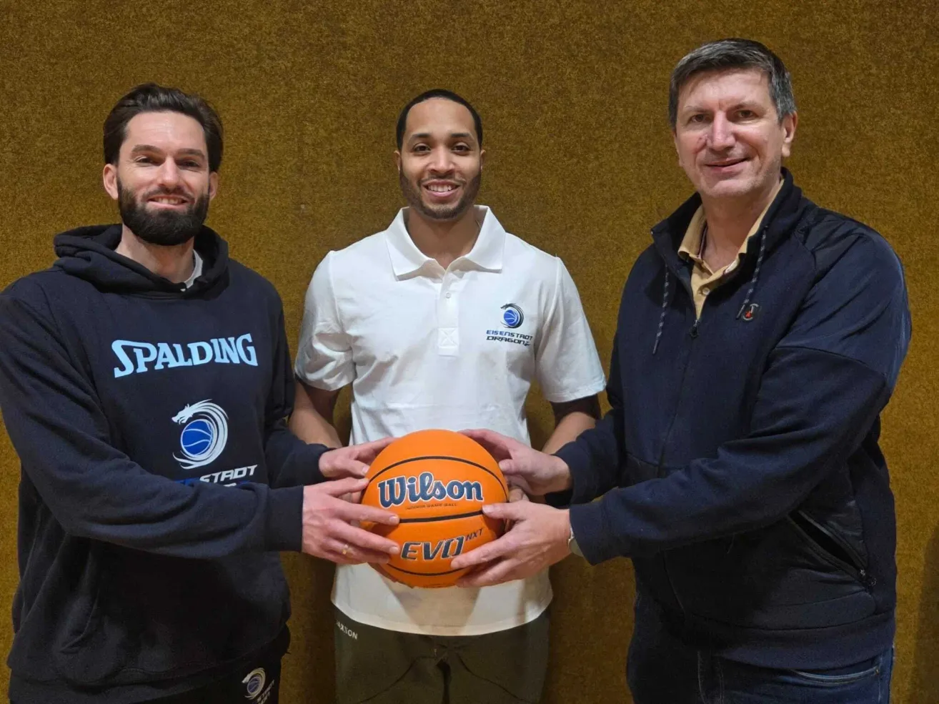 Three men pose for a photo. The man in the middle holds a Wilson basketball. They are all smiling.