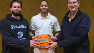 Three men pose for a photo. The man in the middle holds a Wilson basketball. They are all smiling.