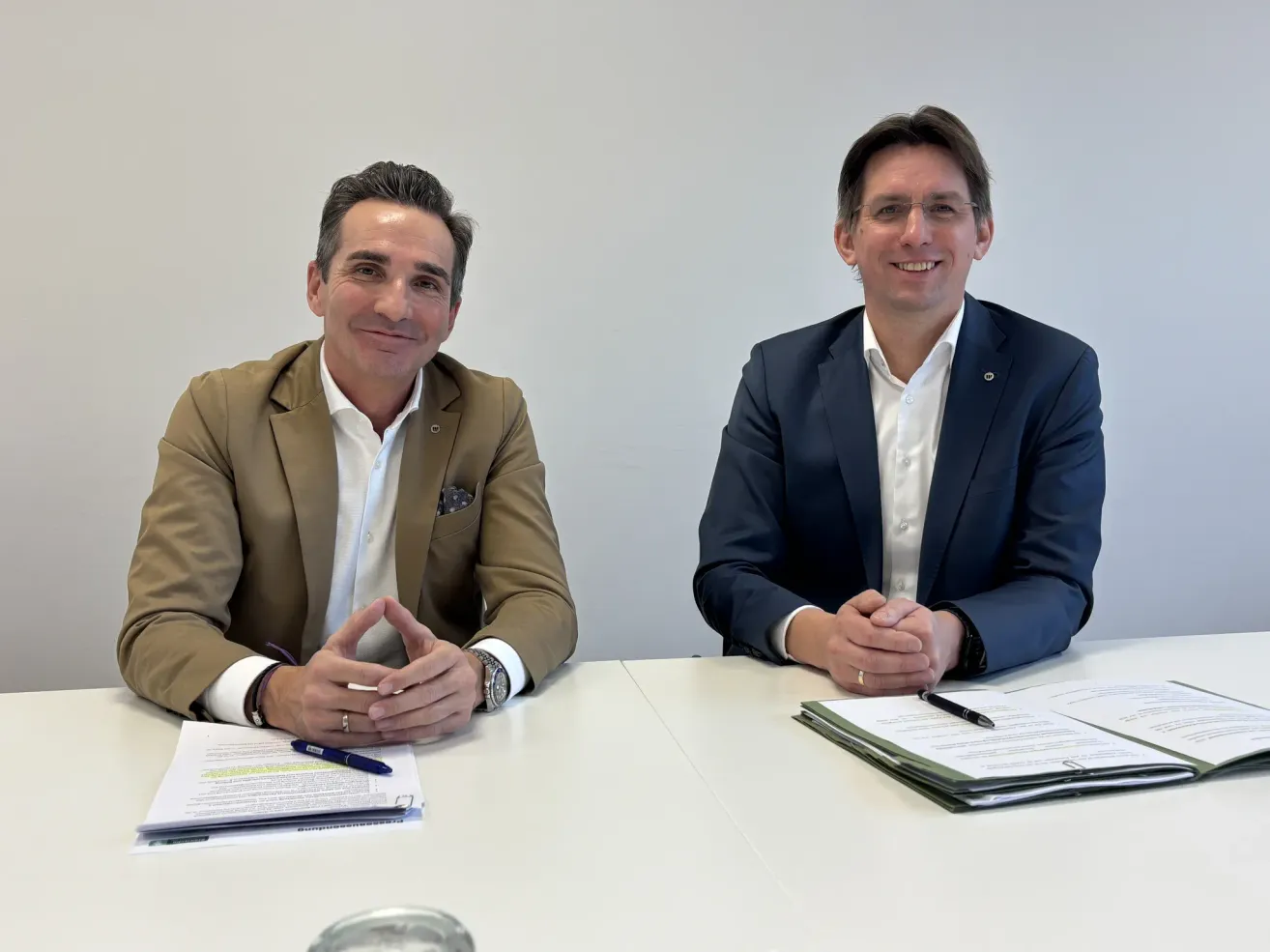 Two men sit at a table, smiling, dressed in business suits, with papers and pens on the table.