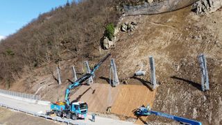 An aerial view of a construction site shows a crane lifting a wooden structure. Workers are present, and metal poles are installed on the slope.