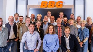 A group of people is posing for a picture in front of a wall with the letters BGG. The people are smiling and wearing casual clothes. Some of them are wearing glasses. The man in the middle is wearing a watch. The woman on the right is wearing a bracelet.