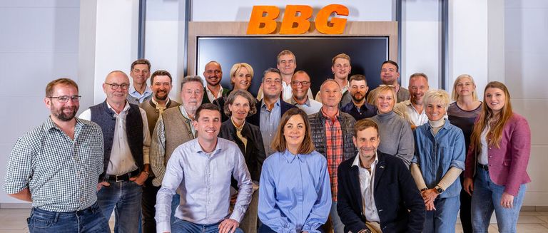 A group of people is posing for a picture in front of a wall with the letters BGG. The people are smiling and wearing casual clothes. Some of them are wearing glasses. The man in the middle is wearing a watch. The woman on the right is wearing a bracelet.