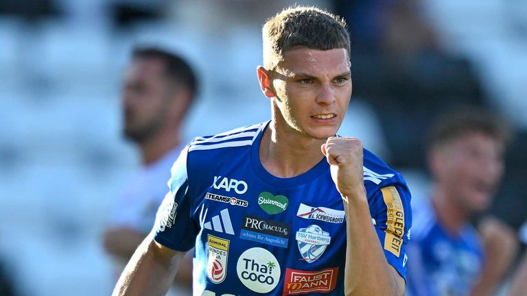 A soccer player in a blue jersey celebrates on the field with another player blurred in the background.