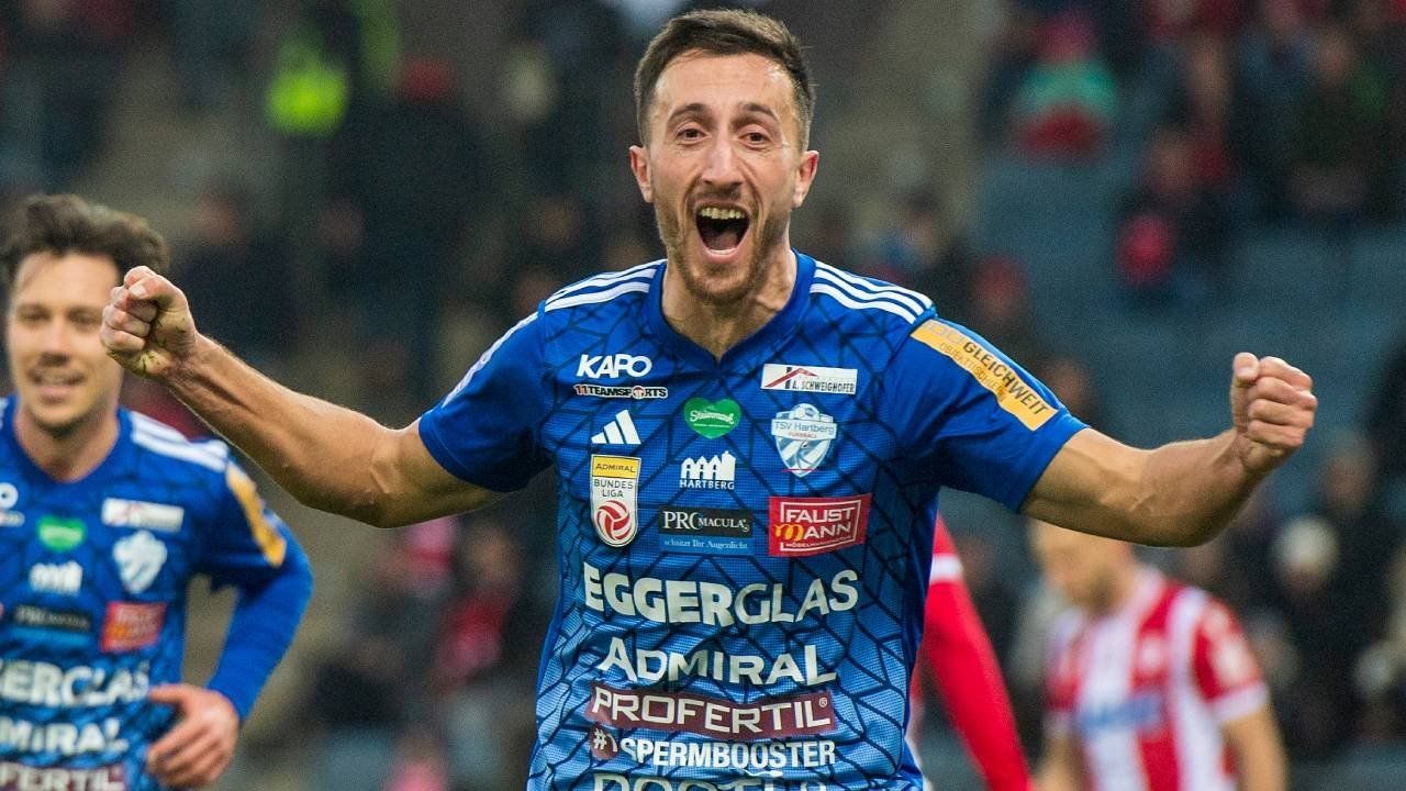 A soccer player celebrates with arms outstretched in a blue jersey with various sponsors' logos.