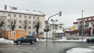 A dark car approaches a traffic light at a snowy intersection. A machinery vehicle is parked behind a fence. Buildings with snow-covered roofs are in the background.