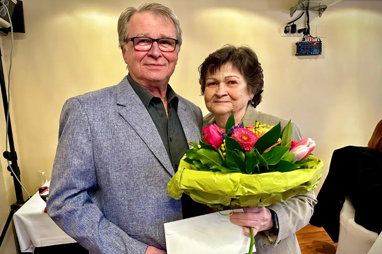 Ein älterer Mann und eine ältere Frau stehen lächelnd zusammen. Der Mann trägt einen grauen Anzug und eine Brille, während die Frau einen Blumenstrauß in der Hand hält. Sie scheinen sich in einem Raum mit einer gelben Wand zu befinden.