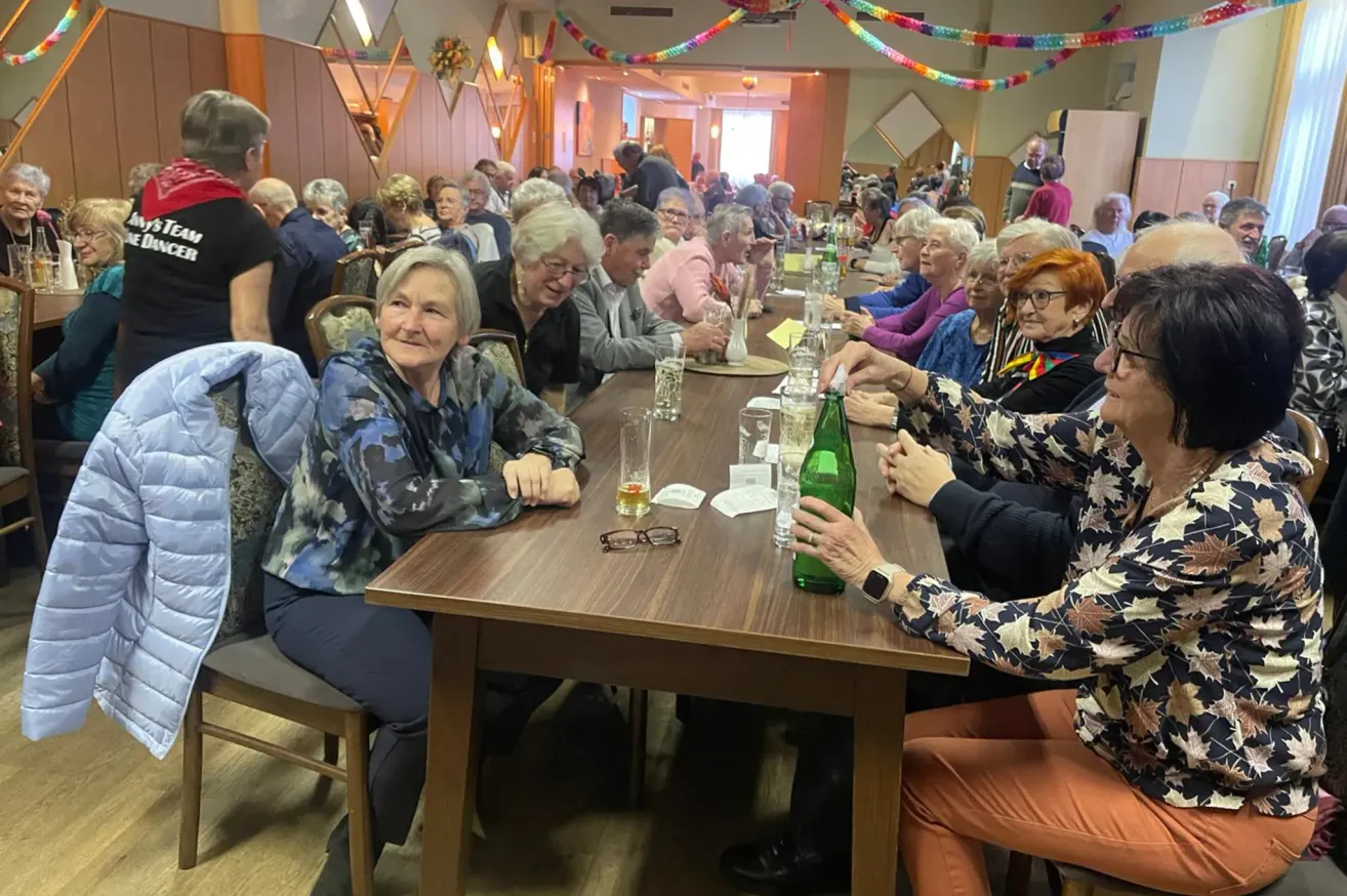 Mehrere ältere Menschen sitzen in einem Restaurant um einen Tisch, lächeln und interagieren. Gläser, eine grüne Flasche und Karten sind auf dem Tisch. Bunte Dekorationen hängen von der Decke herab.