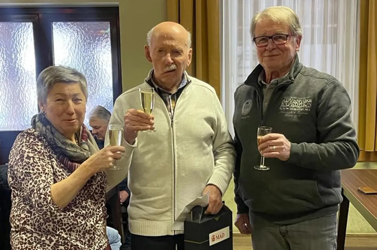 Drei Personen, zwei Männer und eine Frau, stehen eng beieinander und halten Weingläser. Der Mann in der Mitte hält eine Geschenkbox. Dahinter ist ein verschwommenes Bild von Menschen, die auf Stühlen sitzen.