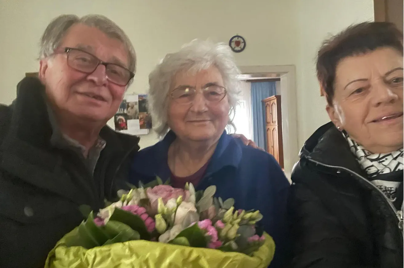 Ein Mann und eine Frau in einer blauen Jacke lächeln und halten einen Blumenstrauß. Eine Frau mit kurzen Haaren steht neben ihnen.