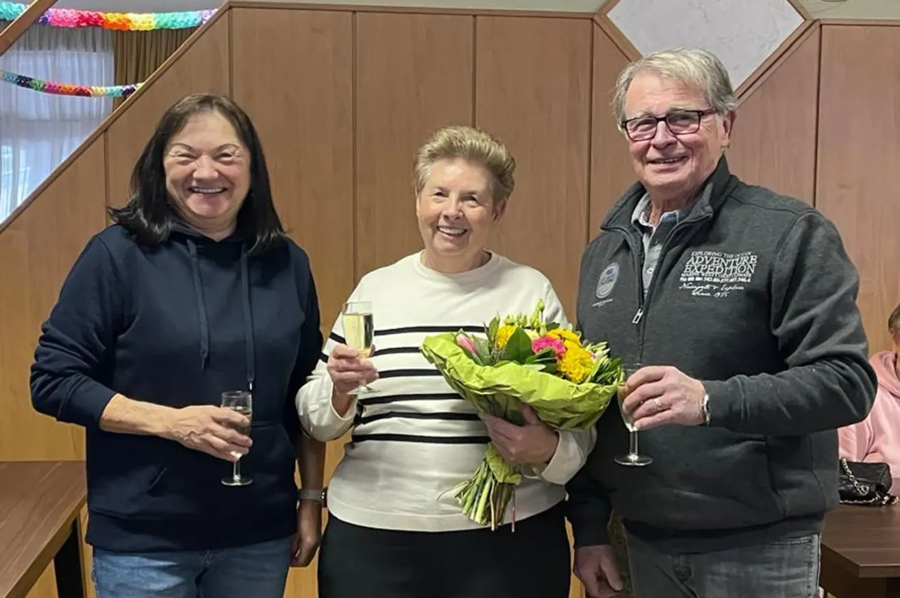 Drei Personen stehen zusammen, lächeln und halten Weingläser und einen Blumenstrauß in den Händen. Eine Frau links trägt eine blaue Kapuzenjacke, während die zentrale Figur, eine Frau, einen Strauß hält und ein weißes Sweatshirt trägt. Der Mann rechts trägt eine Jacke mit 'Adventure Expedition' darauf.