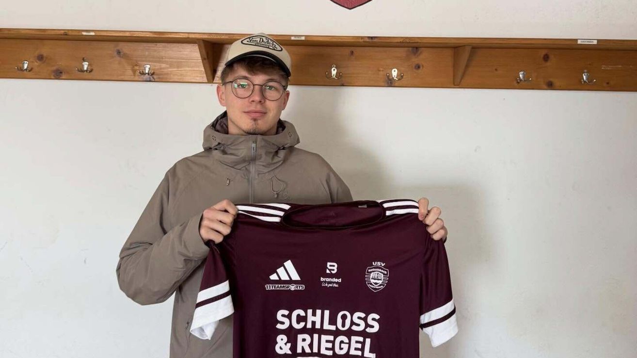 A man in a cap and glasses holds a maroon jersey with white stripes, displaying Schloss & Riegel on the front. He stands in front of a white wall.