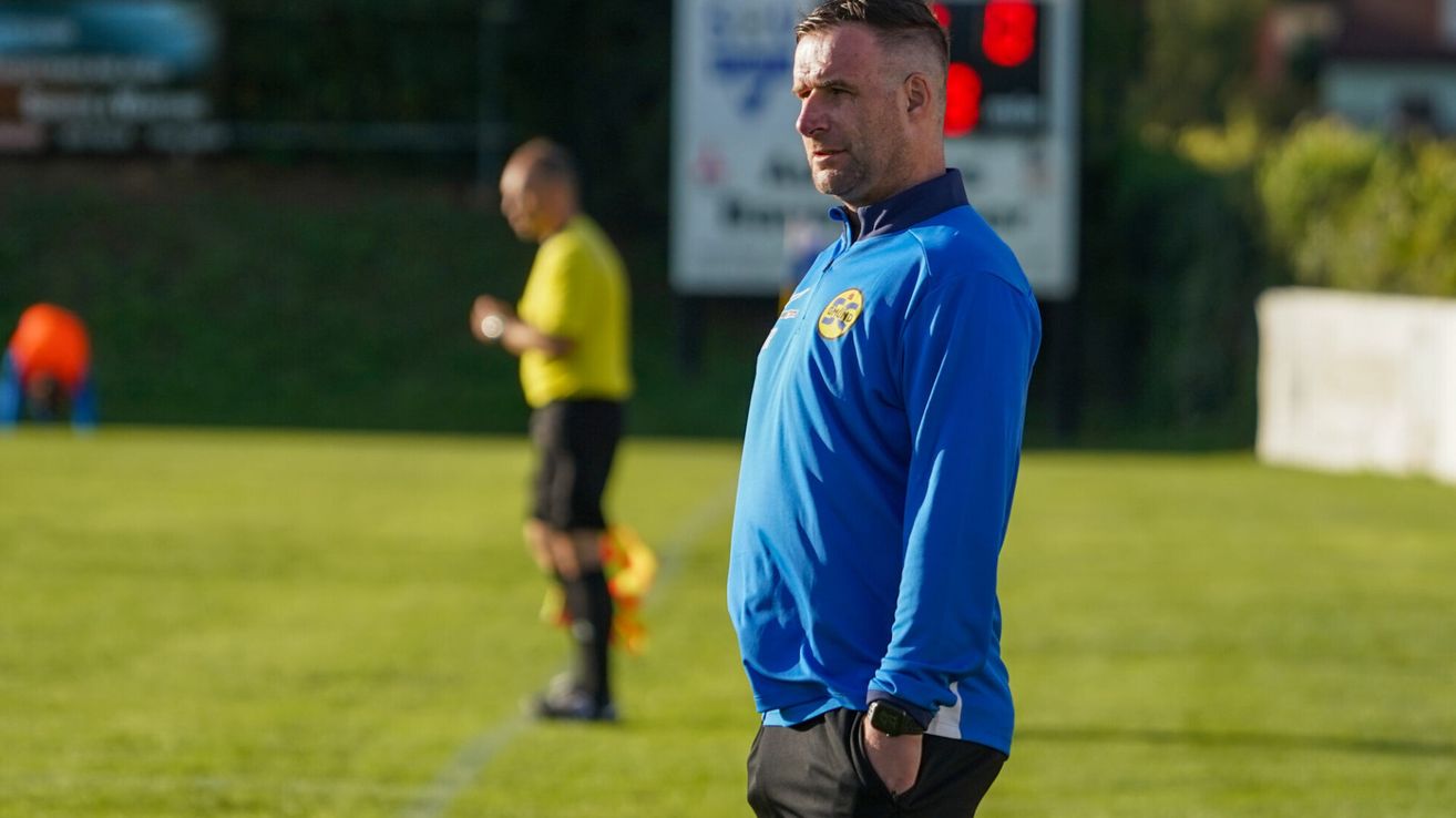 A man wearing a blue shirt with a logo stands on a soccer field, looking serious, while another man in a yellow shirt walks behind him. A digital scoreboard is visible in the background.