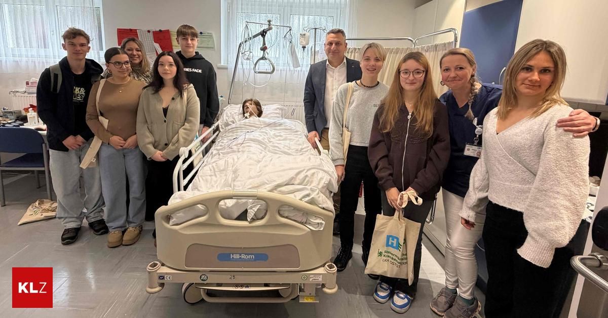 A group of people, including a man in a suit, are standing around a hospital bed, posing for a photo. The bed has a mannequin on it, and a woman is holding a bag.