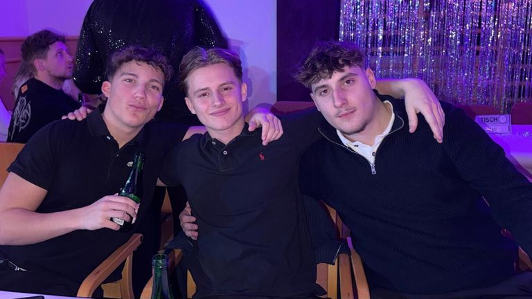 Drei junge Männer posieren für ein Foto. Der mittlere Mann trägt ein schwarzes Poloshirt. Der Mann auf der rechten Seite hat seinen Arm um den mittleren Mann gelegt. Sie lächeln und scheinen auf einer Party zu sein.