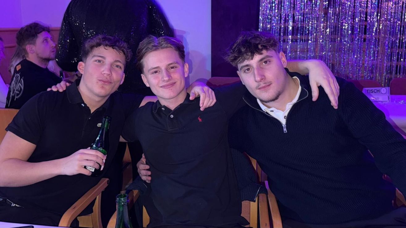 Drei junge Männer posieren für ein Foto. Der mittlere Mann trägt ein schwarzes Poloshirt. Der Mann auf der rechten Seite hat seinen Arm um den mittleren Mann gelegt. Sie lächeln und scheinen auf einer Party zu sein.