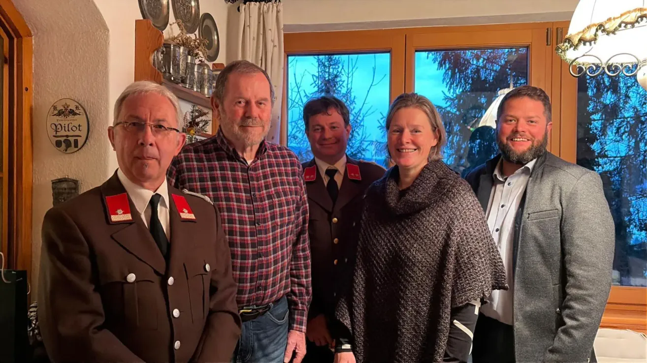 Fünf Personen stehen in einem Raum zusammen und lächeln für ein Foto. Ein Mann in Uniform steht links, und eine Frau in einem gestrickten Cape steht rechts. Dahinter tragen zwei Männer Uniformen mit Abzeichen. Der Hintergrund zeigt ein Fenster mit Bäumen.