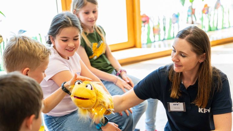Eine Frau spielt mit einer Puppe, während zwei junge Mädchen hinter ihr sitzen. Ein Mädchen lächelt und schaut die Frau an. Die Frau hat einen Namensschild mit dem Text KFV und Veronika.