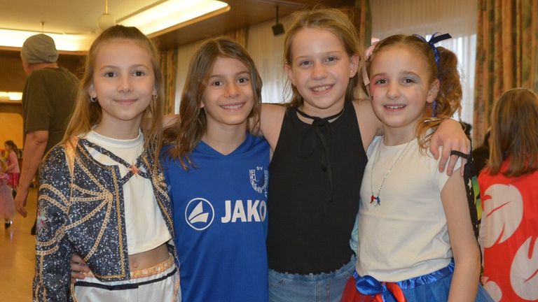 Four young girls are smiling for a photo. The girl in the middle wears a blue shirt with a white logo. The girl on the right has a pink bow in her hair.