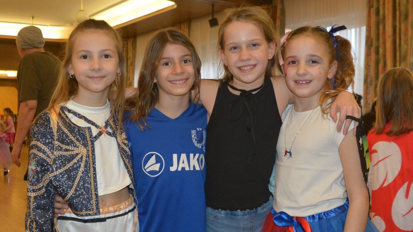 Four young girls are smiling for a photo. The girl in the middle wears a blue shirt with a white logo. The girl on the right has a pink bow in her hair.