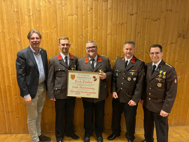 Five men in uniforms stand in front of a wooden wall. They hold a framed certificate for Erich Dunder, signed by the mayor.