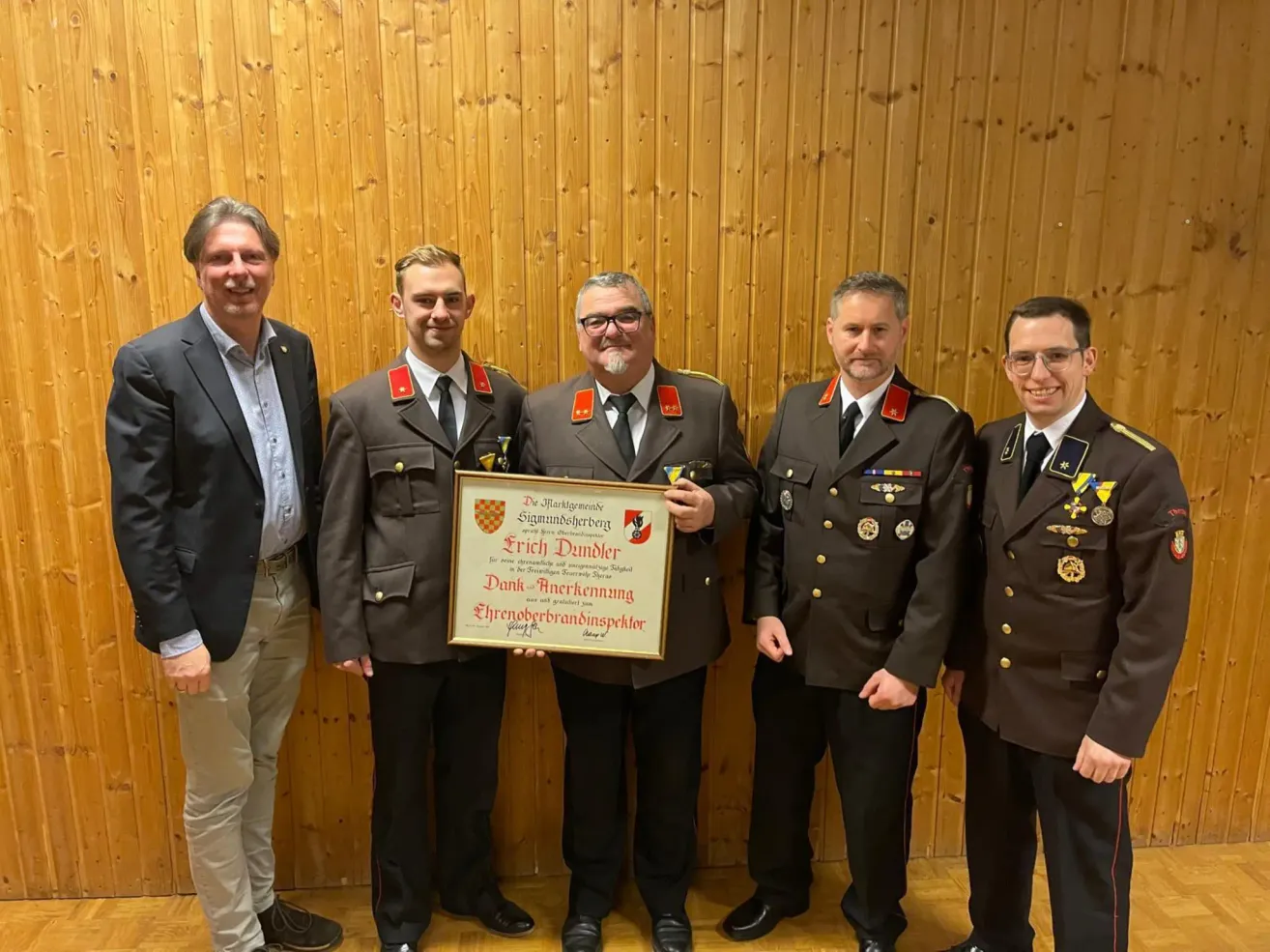 Five men in uniforms stand in front of a wooden wall. They hold a framed certificate for Erich Dunder, signed by the mayor.