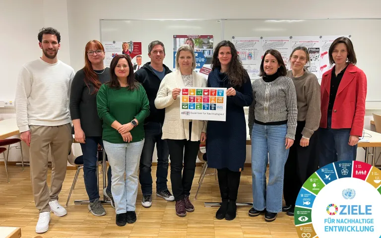 Eine Gruppe von Menschen steht in einem Raum, lächelt und eine hält ein Poster mit Symbolen und Text. Hinter ihnen befinden sich Poster an der Wand.