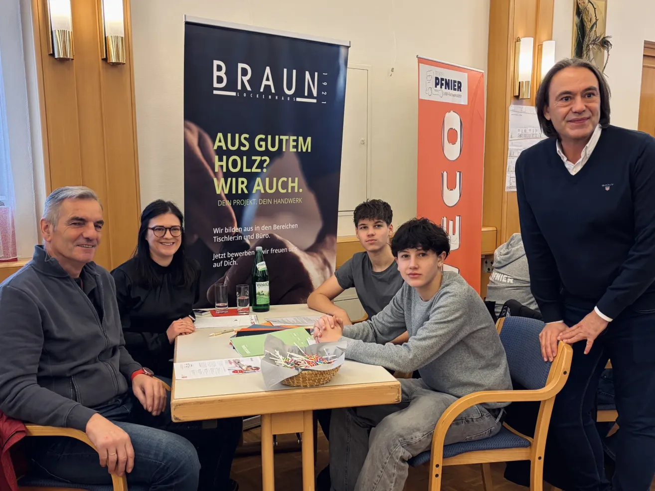 Eine Gruppe von Menschen sitzt um einen Tisch herum, vor ihnen ein Banner, das für Braun Lockenhaus wirbt. Ein Mann und eine Frau lächeln und posieren für ein Foto.