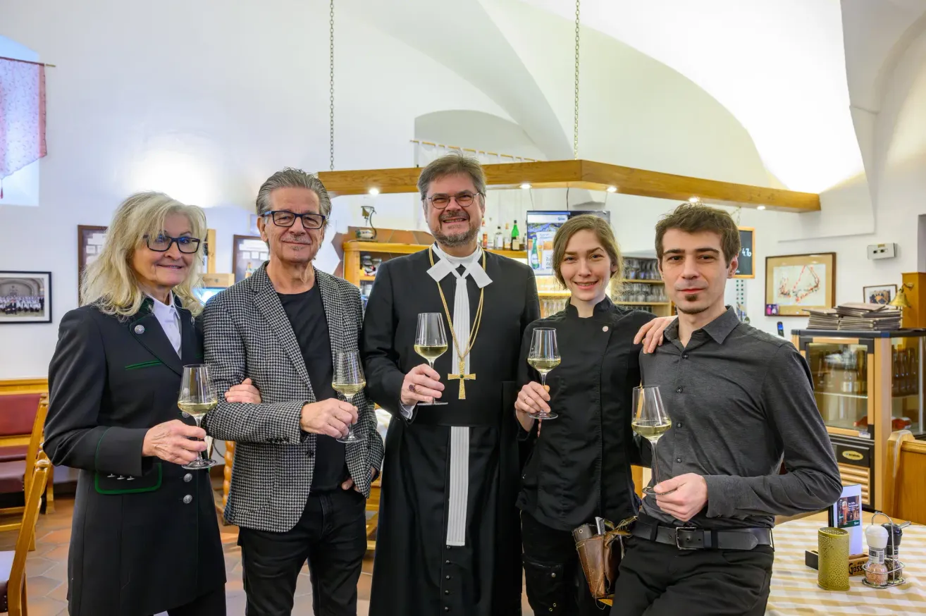 Five people are standing and smiling, holding wine glasses, possibly in a restaurant. They are dressed formally, with the man in the middle wearing a priest's attire.