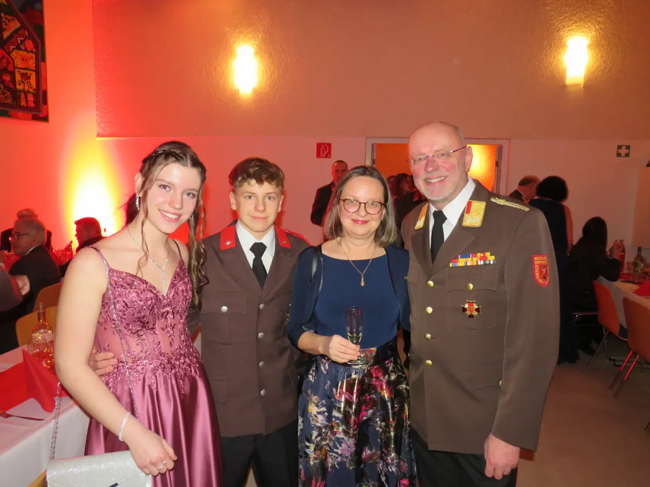 Vier Personen posieren für ein Foto. Ein Mann in Uniform, eine Frau mit einem Weinglas, ein Mädchen und ein Junge, alle lächeln. Hinter ihnen stehen weitere Personen. Eine rote Wand und eine beleuchtete Lampe sind im Hintergrund.