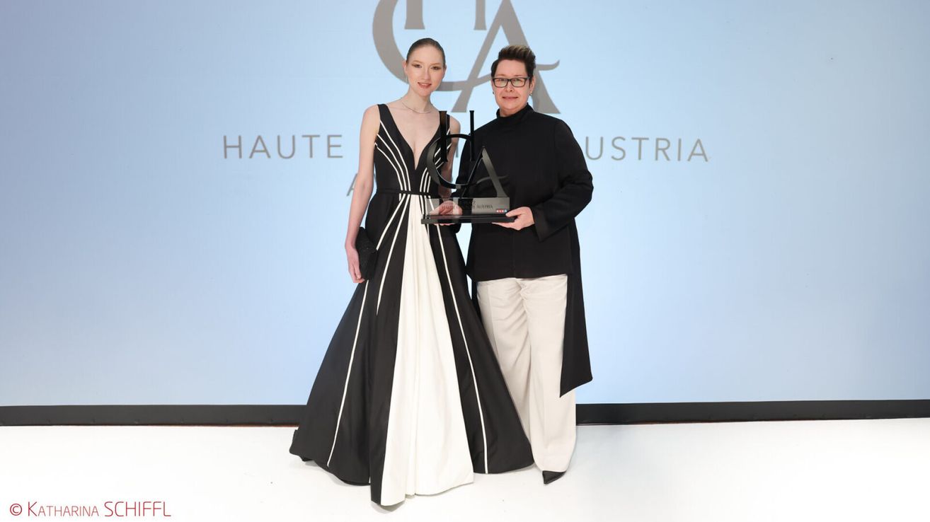 Two individuals stand next to each other, holding a trophy. The woman wears a black and white gown and the man wears a black coat. Behind them, a blue backdrop displays the text 'HAUTE AUSTRIA.'