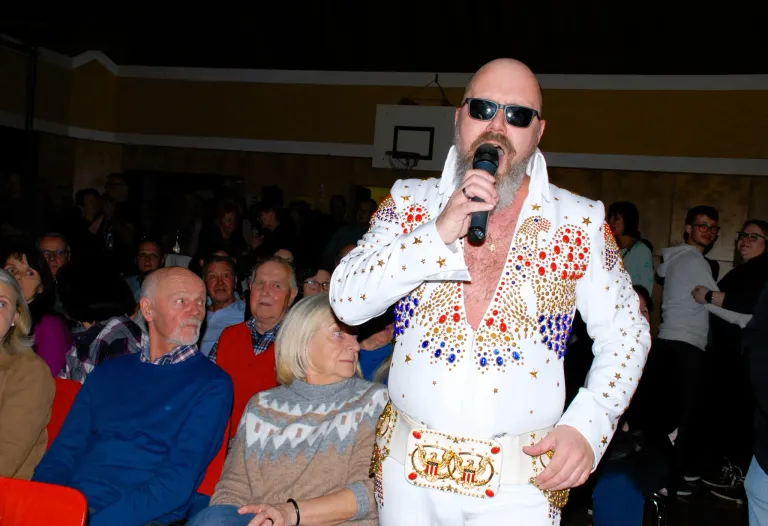 Elvis-Imitator singt mit einem Mikrofon in einem überfüllten Raum, zwei ältere Menschen sitzen vor ihm und schauen zu.