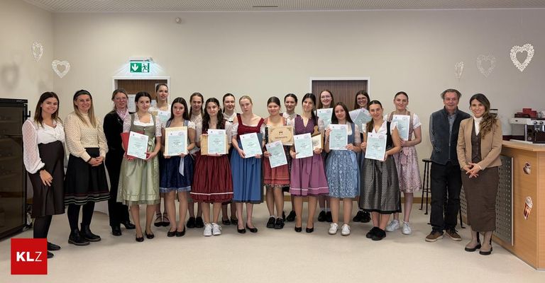 A group of young women in traditional dresses pose for a photo while holding certificates in a room with white walls and a wooden door.