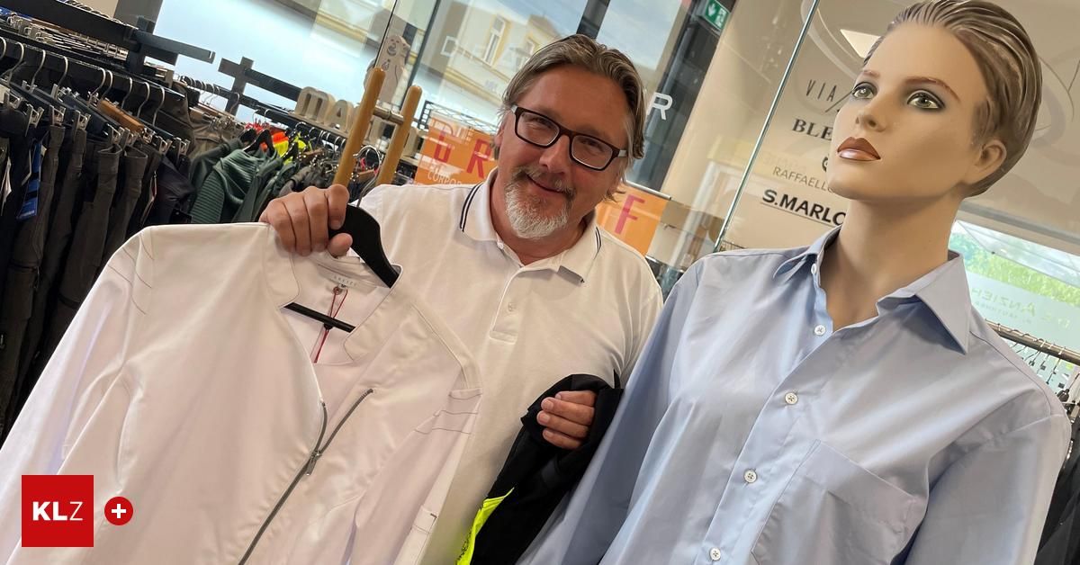 A man with glasses holds a white jacket and poses next to a mannequin wearing a blue shirt inside a clothing store.