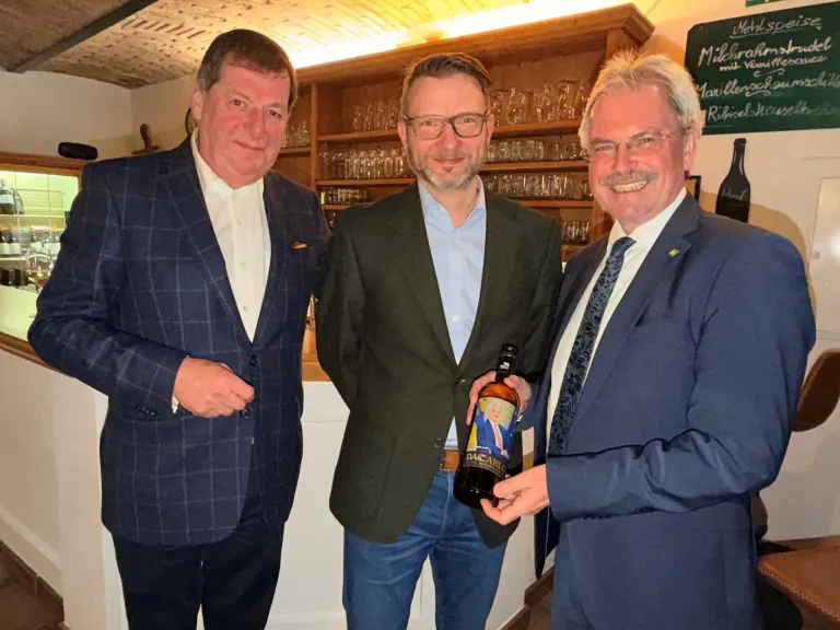 Three men in suits stand next to each other in a bar. The man in the middle holds a bottle labeled 'DACARO'. They all smile for a photo.