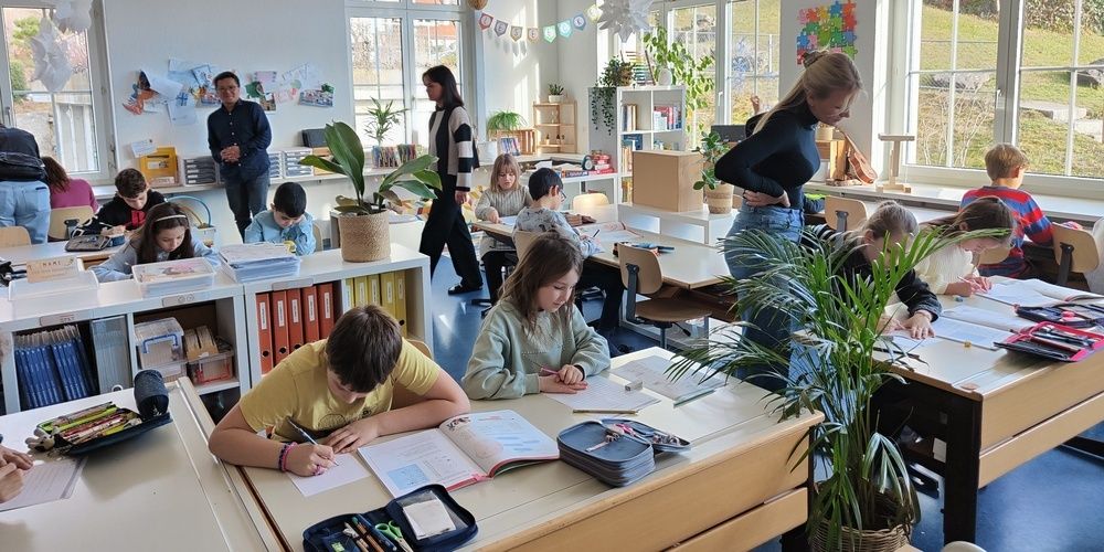 A classroom with children sitting at desks, one boy writes on paper while another looks at a book. A teacher stands near a woman, and plants and books are around.