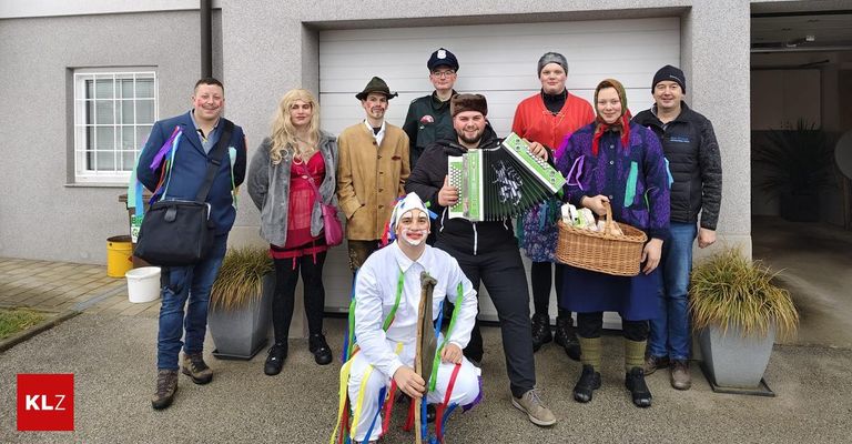Eine Gruppe von Menschen in Kostümen posiert für ein Foto. Eine Person in einem weißen Kostüm hält ein Akkordeon. Eine andere Person hält einen Korb. Die Leute stehen vor einem Gebäude.