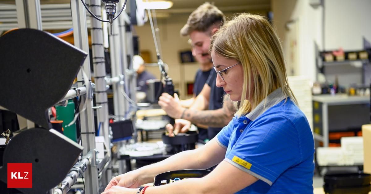 A woman in a blue shirt works on a machine in a factory, while two men, one with a tattoo, work behind her. The area is filled with machinery and tools.