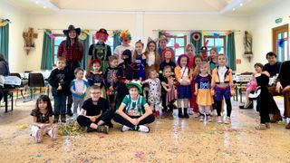 Eine Gruppe von Kindern und Erwachsenen in Kostümen posiert für ein Foto in einem Raum mit Konfetti auf dem Boden. Einige tragen Clown-, Skelett- und Feuerwehrkostüme. Ein Mann in einem Cowboyhut steht neben einer Frau in einem rot karierten Kleid.