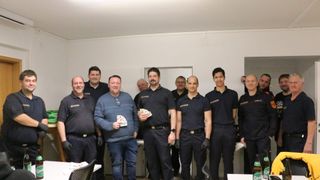 Eine Gruppe von Männern in Uniformen und mit Karten in den Händen, die für ein Foto lächeln. Der Mann in der Mitte hält ein Kartenspiel, und sie stehen vor einer weißen Wand.