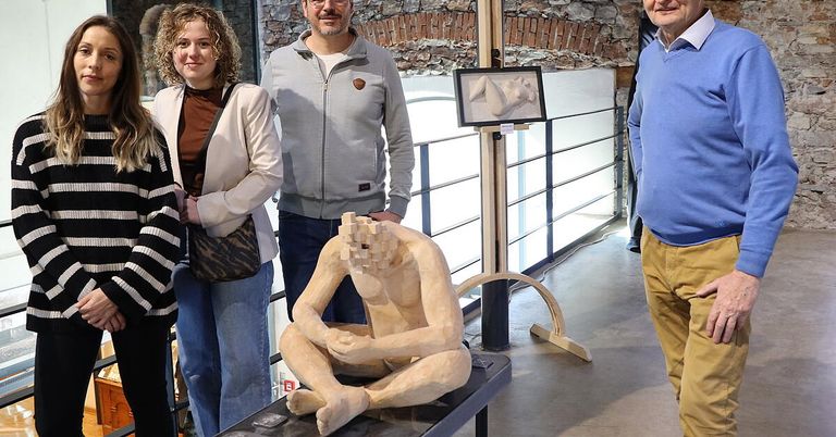 Drei Personen stehen vor einer Ziegelwand, ein Mann in der Mitte sitzt auf einer Plattform. Sie alle lächeln.