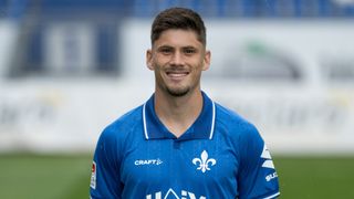 A man wearing a blue polo shirt with a logo on the left chest, smiling and standing in an outdoor area. The shirt features white trims.