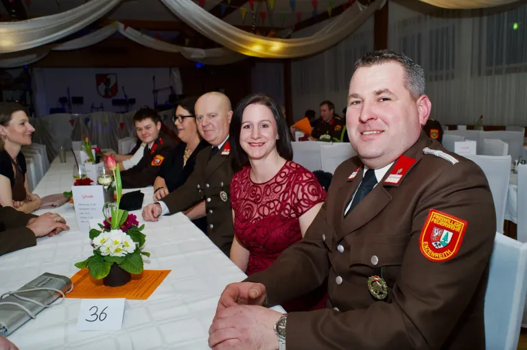 Ein lächelnder Mann in Uniform sitzt an einem Banketttisch mit einer Frau in einem roten Kleid. Dahinter sitzen eine Gruppe uniformierter Personen, und ein geschmückter Tisch steht im Vordergrund.